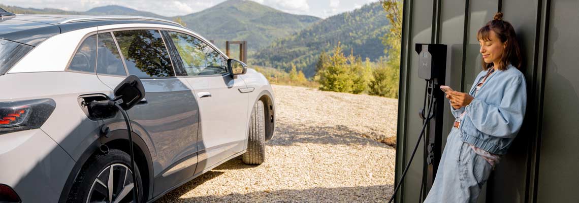 Woman stands with a smartphone and waits for electric car to be charged near a charging station at home