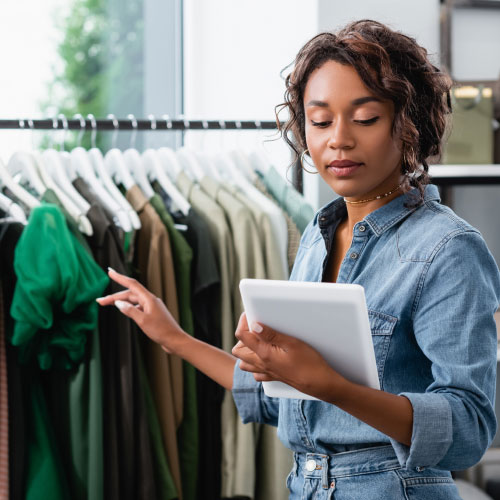 young saleswoman holding digital tablet and pointing at clothing on rack