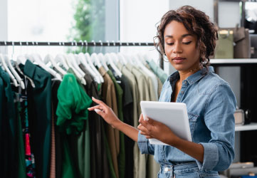 young saleswoman holding digital tablet and pointing at clothing on rack