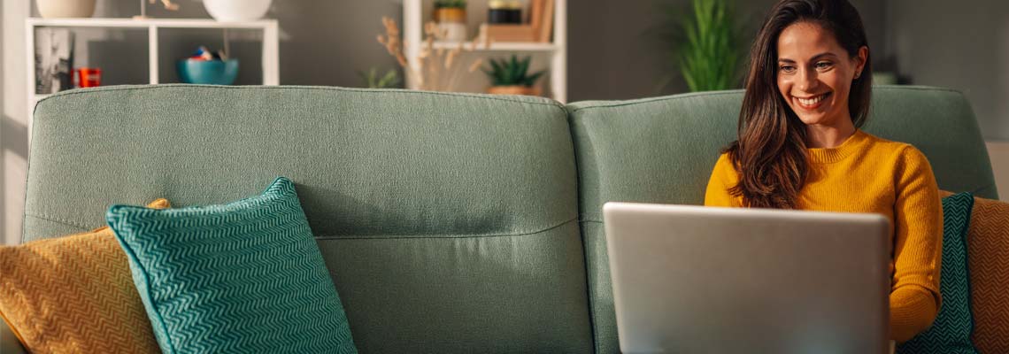 A cheerful woman is sitting at her home on couch and using a laptop