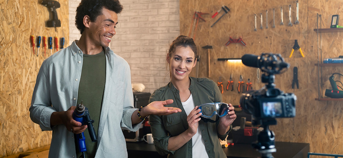 2 people in a workshop talking to a SLR camera and holding safety goggles