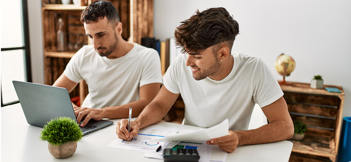 two men casually dressed reviewing finances at a laptop. One is using the laptop while the other is using a calculator with paperwork scattered across the desk