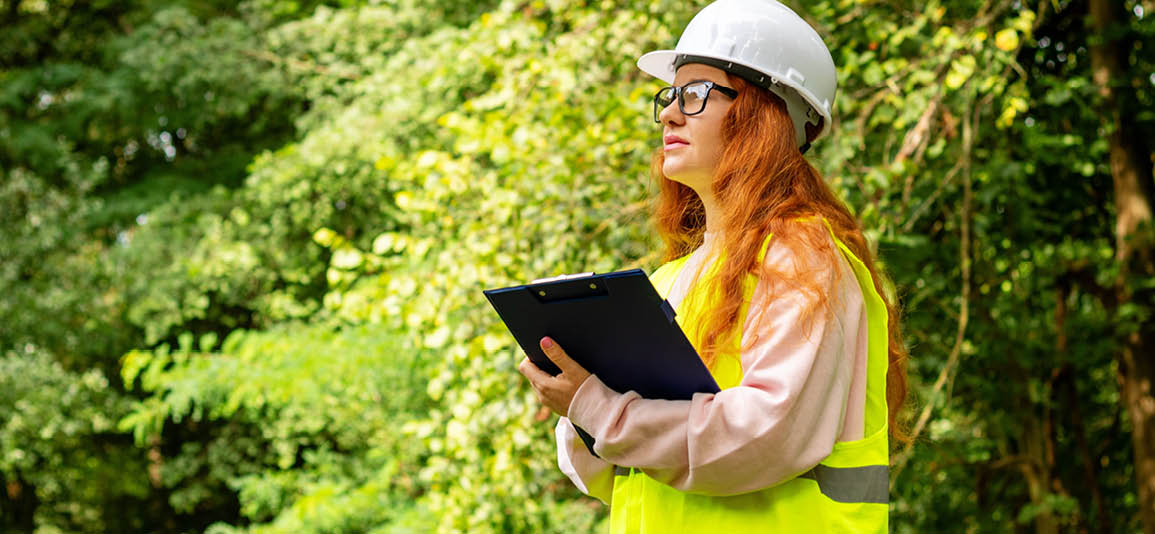 Woman wearing a hard hat in a forest, holding a clipboard reviewing safety