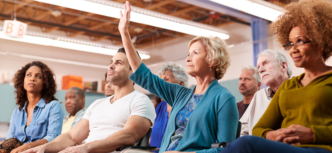 Women sitting in the front row of a neighbourhood meeting raising her hand to ask a question.