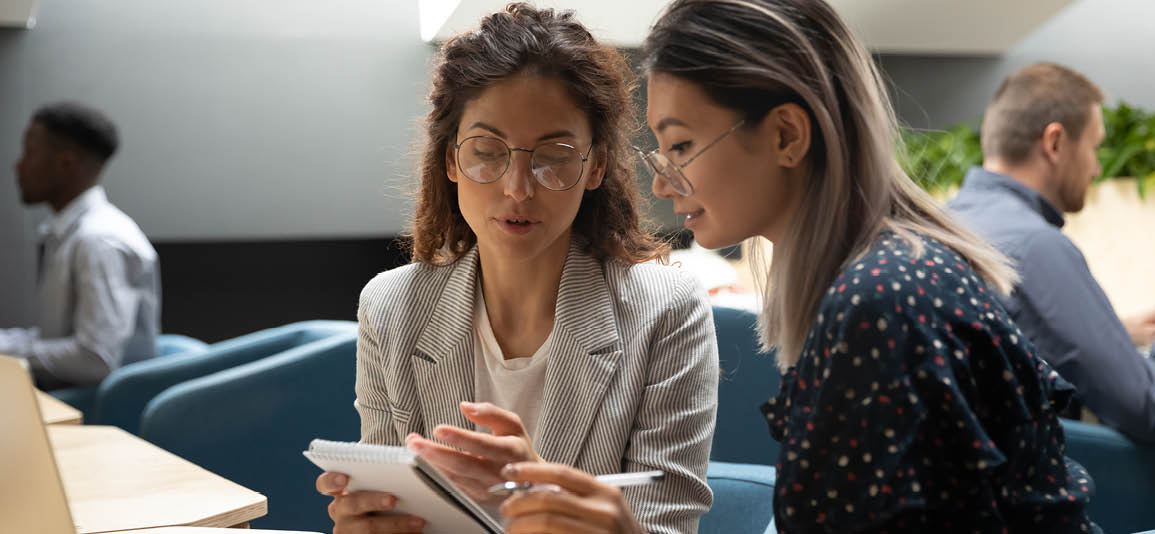 two women working in an office environment using a laptop and writing down notes on a notepad