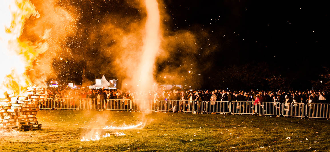 Bonfire on a field with audience behind fencing at a safe distance away