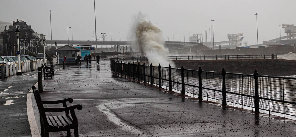 Stormy coastal scene with large waves crashing over a seawall onto a wet promenade. Empty benches line the walkway, and metal railings separate the path from the turbulent water. The sky is overcast, and visibility is reduced by rain and mist