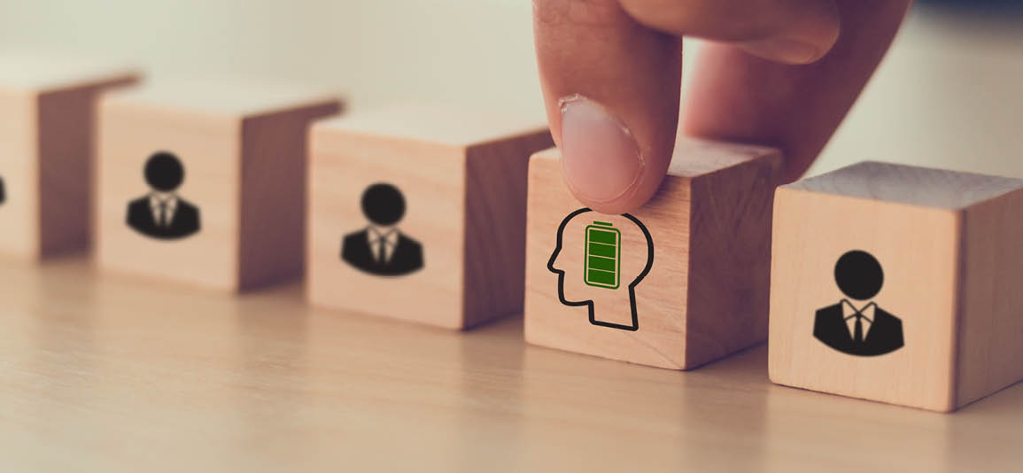 A row of wooden blocks featuring icons of people, with one block highlighted showing a silhouette of a head and a battery symbol inside, representing mental energy and wellbeing.