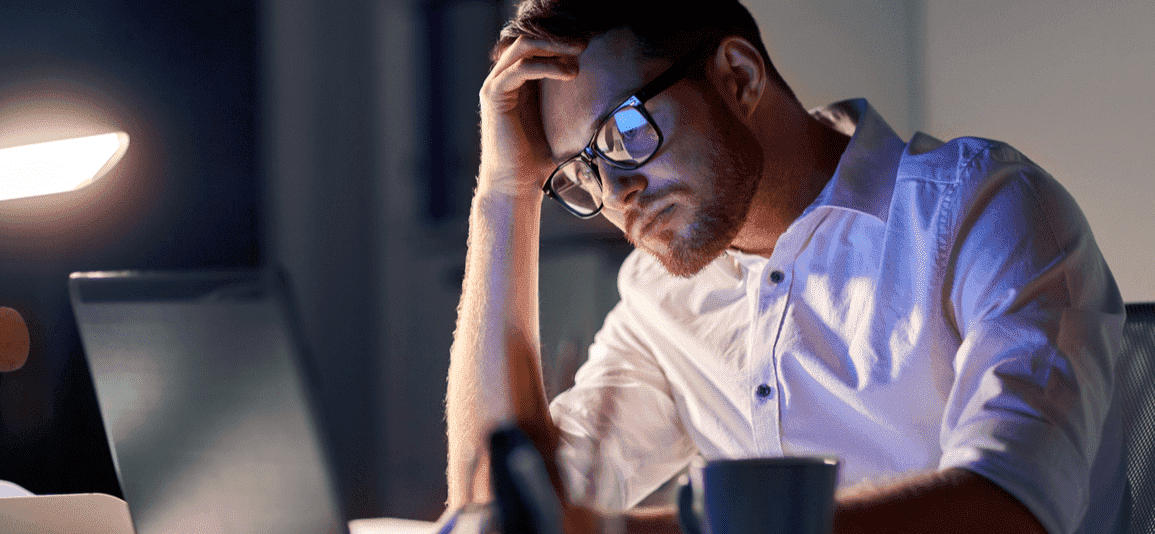Man in white shirt and glasses in front of laptop screen looking worried with his head in his left hand 