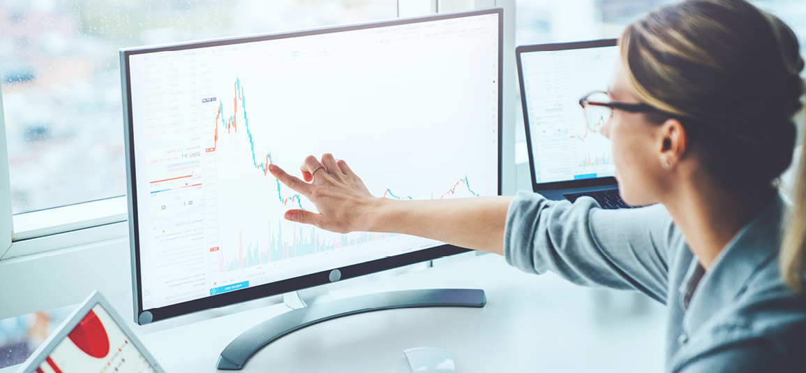 Back view of business woman sitting at desktop front PC computer with financial graphs and statistics on monitor.