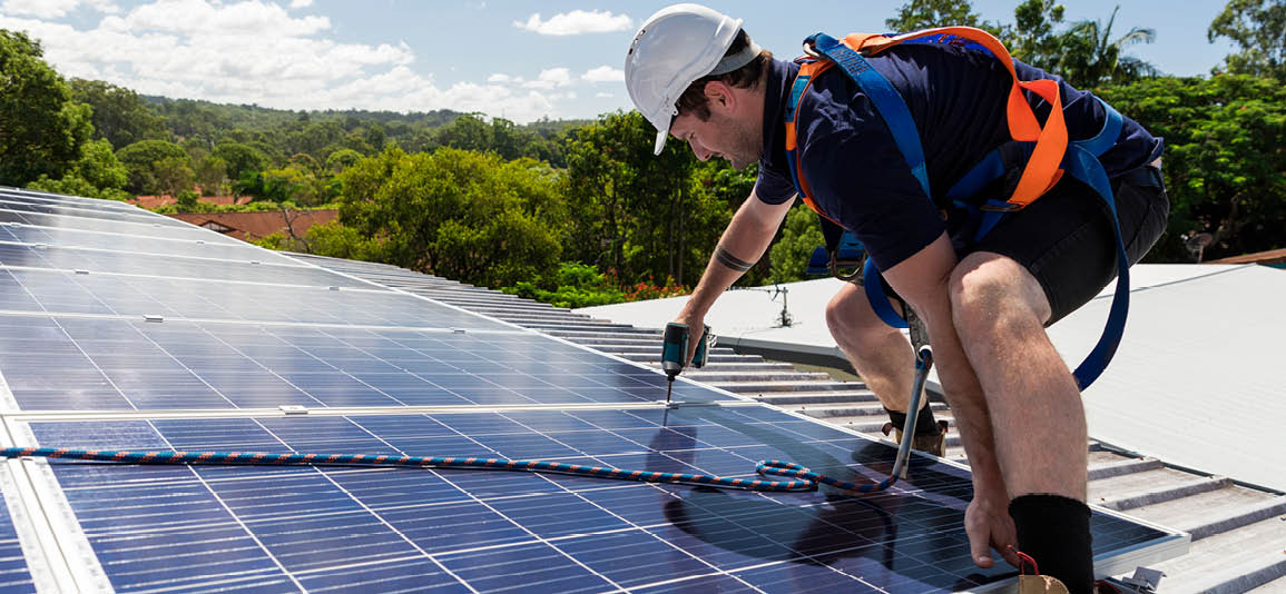 Solar panel technician drilling solar panel while tethered to a roof, wearing a hard hat and safety boots