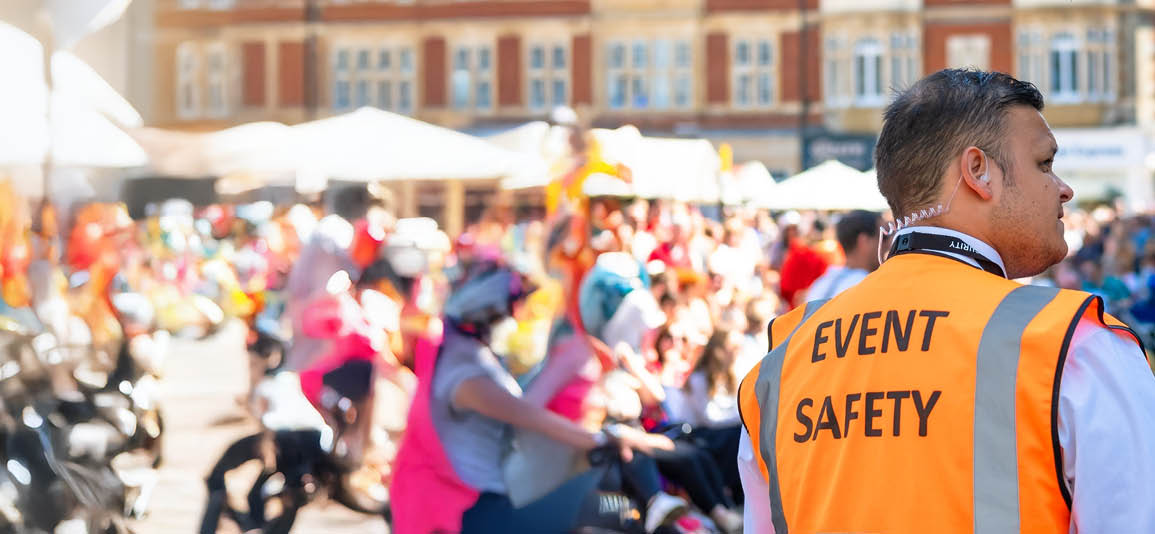 Event safety staff member wearing a high‑visibility vest overseeing a busy outdoor public gathering in a town square.