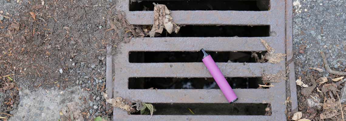 A purple e-cigarette vape has been discarded and left lying on a metal water drain cover